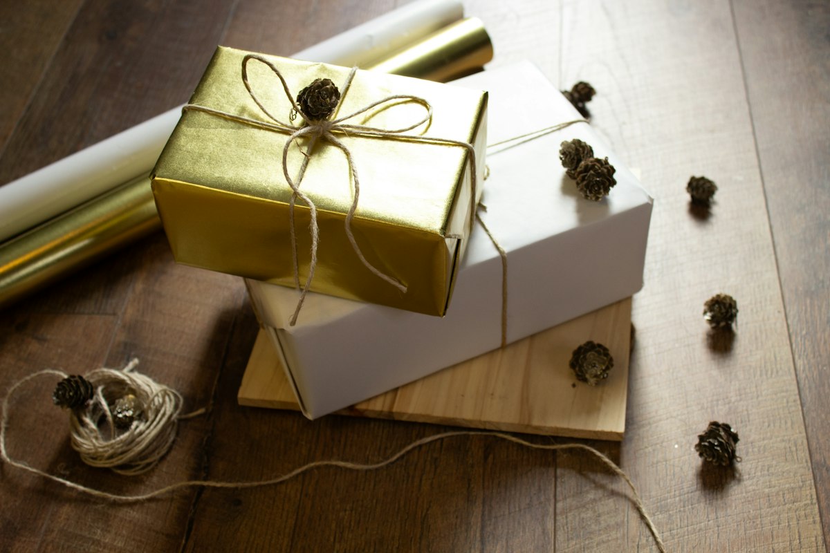 Gift boxes wrapped in gold and white paper, tied with twine and set on a wooden floor beside a roll of gold wrapping paper